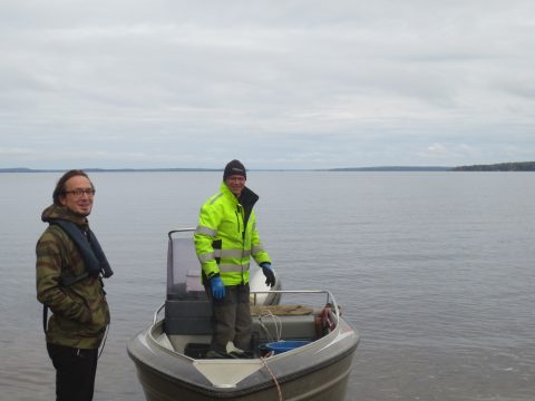 Mies seisoo rannassa veneen vierellä. Toinen mies seisoo veneessä, taustallaan järvenselkä.