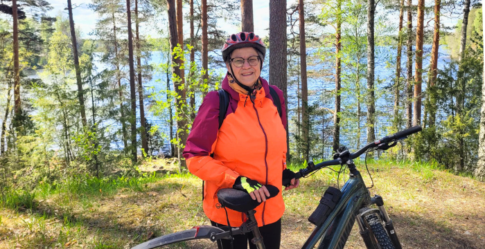Minna Uosukainen seisoo polkupyörän kanssa metsäpolulla, taustalla vesistö.