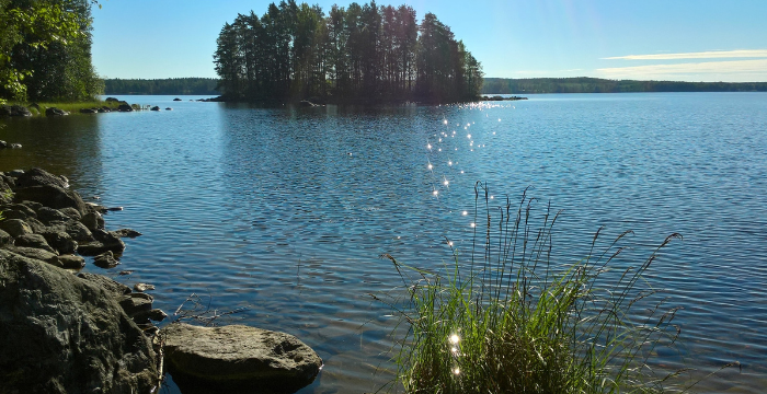 Aurinkoinen järvimaisema, etualalla rantakiviä, taustalla pienehkö saari.