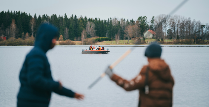 Nuoria ongella järven rannassa, taustalla vene, jossa ihmisiä kalastamassa.